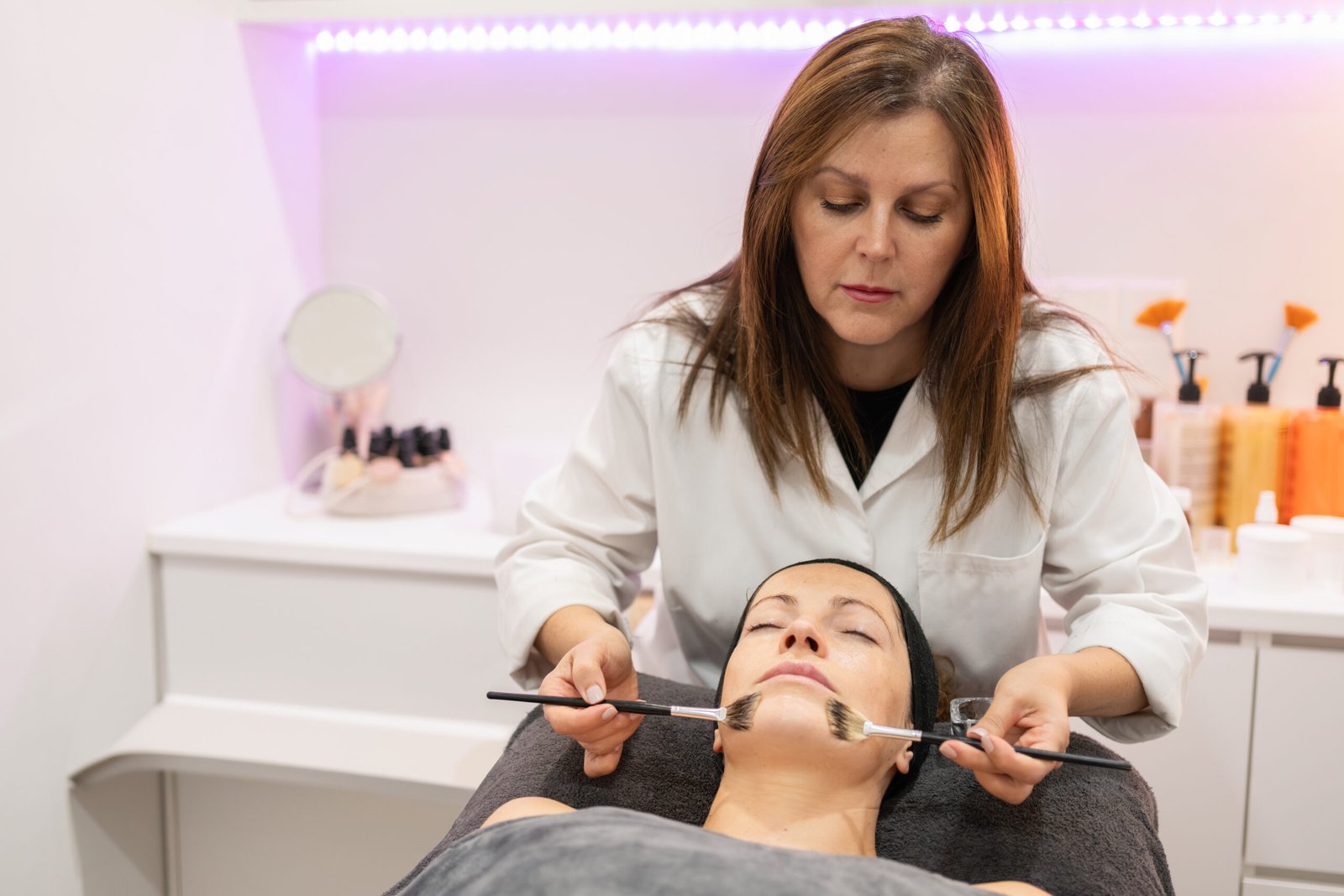 Concentrated brunette beautician applying facial cleansing oil with brush on woman lying face up with eyes closed during cosmetology session in beauty salon against purple lights in background with cosmetics on counter